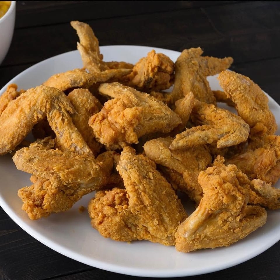Plate piled with golden-brown fried chicken wings on a dark wooden table, with a bowl of dipping sauce partially visible.