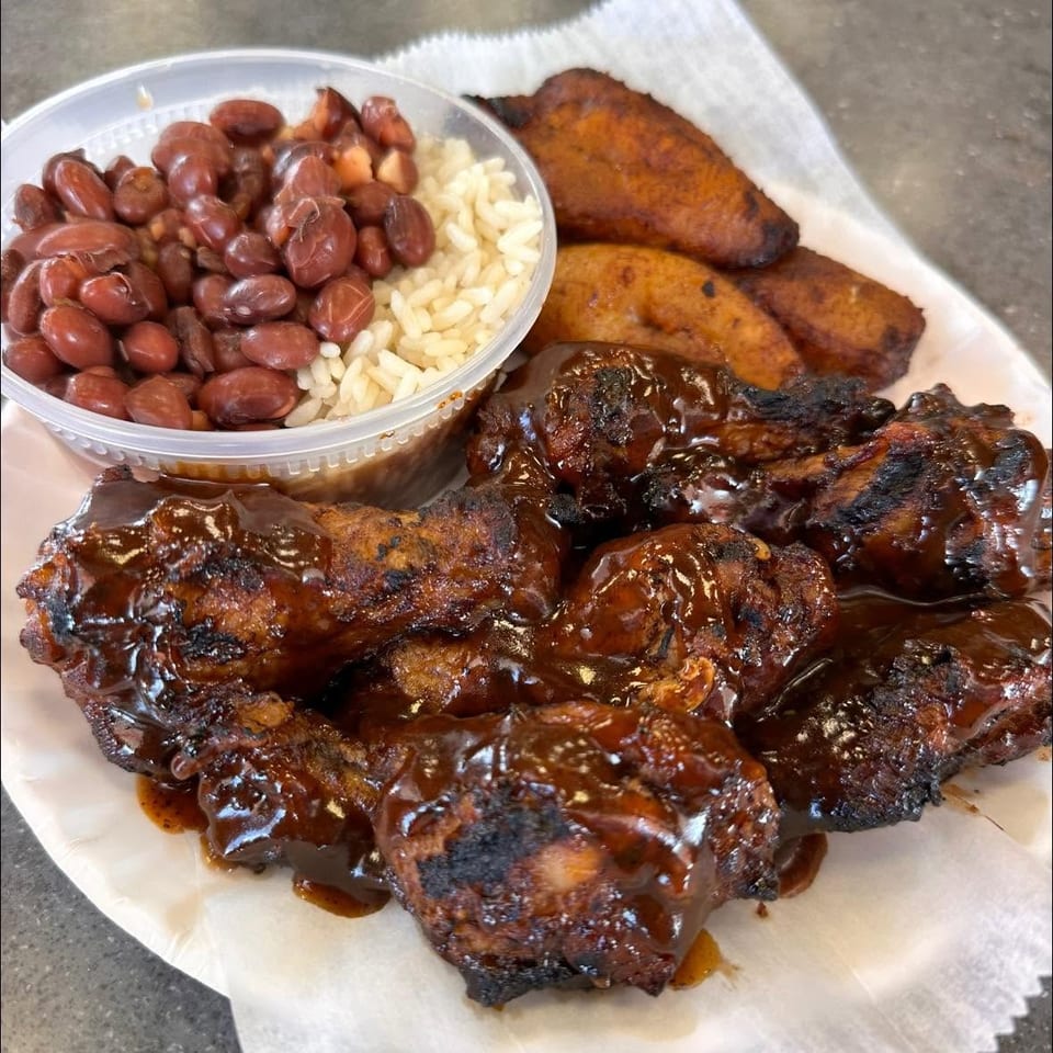 Plate of glazed grilled chicken wings with fried plantains and a container of red beans and rice.