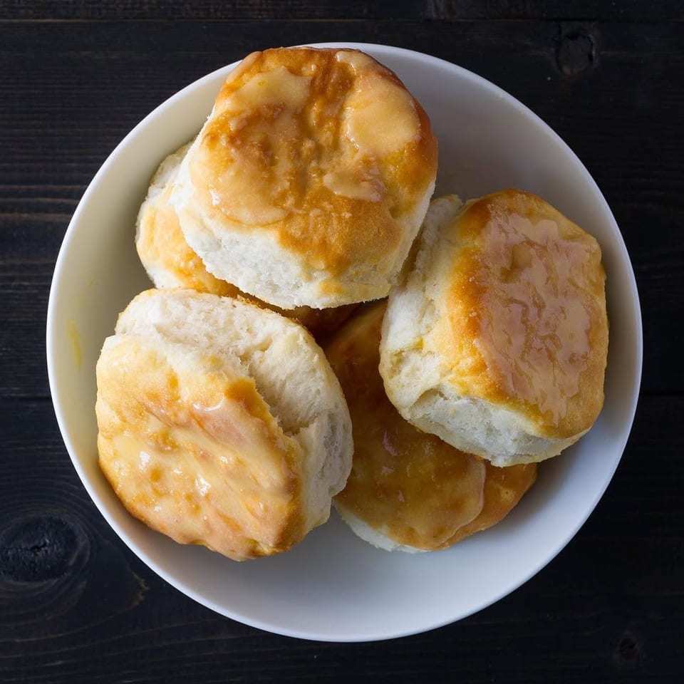 Several golden-glazed biscuits arranged in a white bowl on a dark wooden table.
